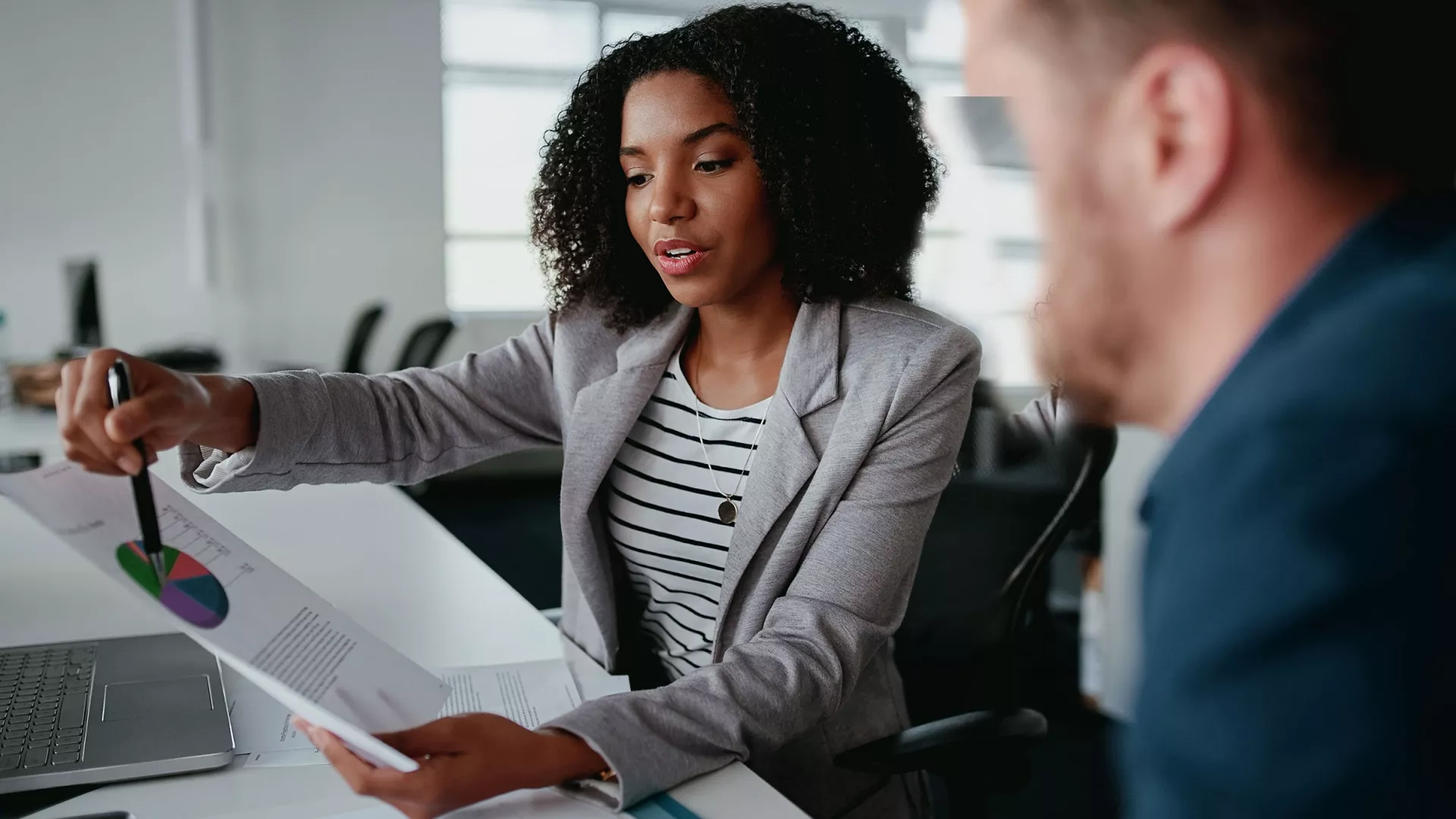 Confident young businesswoman holding document showing increasing statistic to partner in office